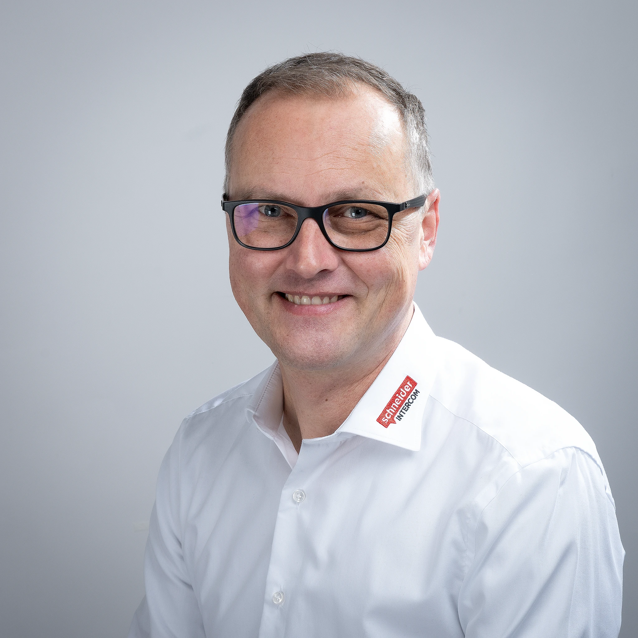 Lächelnder Mann mit Brille und Schneider Intercom Logo auf weißem Hemd.