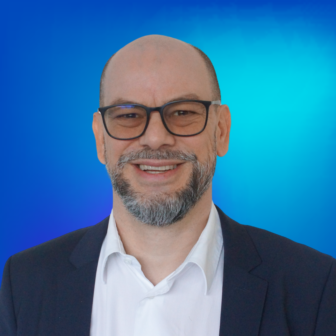 Lächelnder Mann mit Brille und Bart vor blauem Hintergrund.