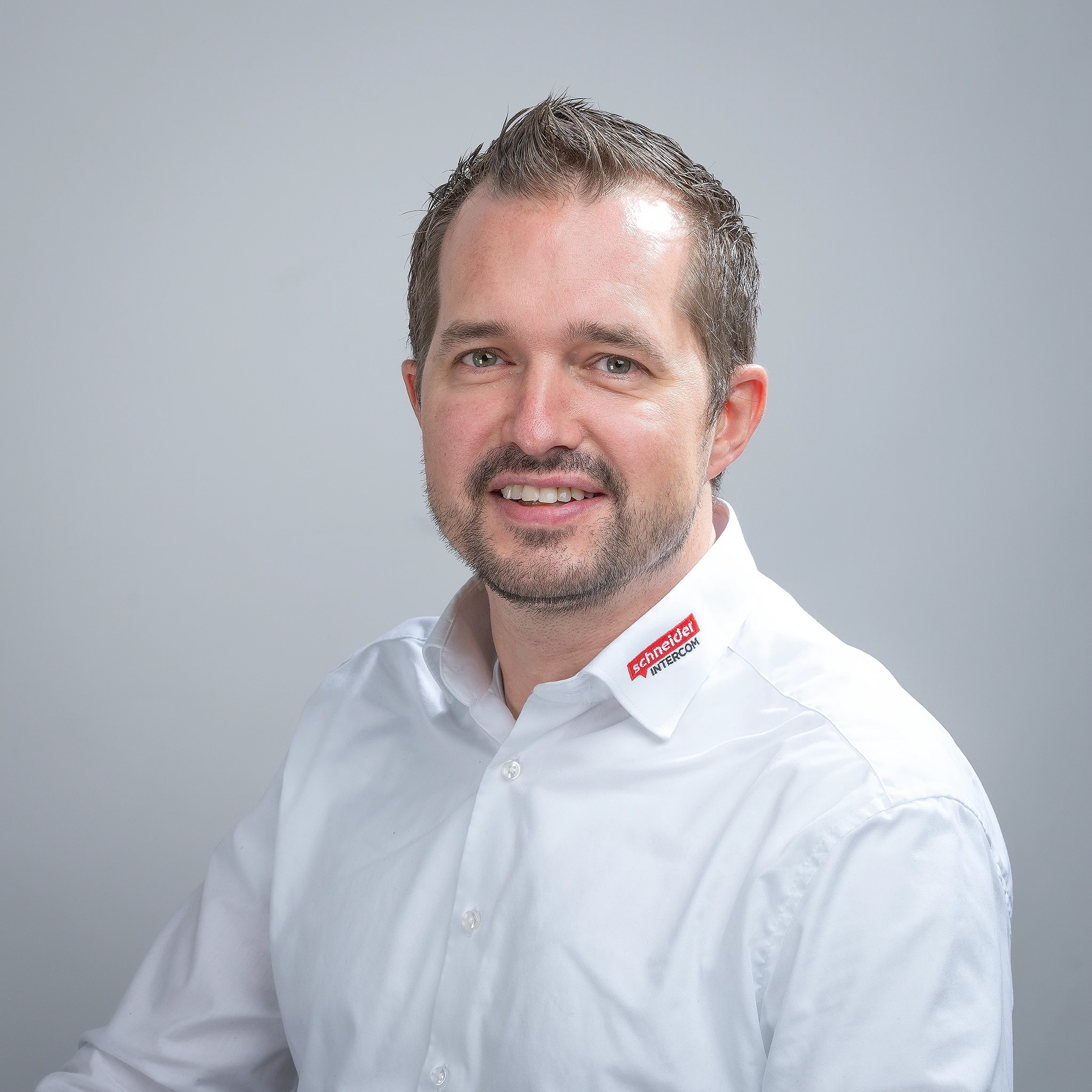 Lächelnder Mann im weißen Hemd mit "Schneider INTERCOM" Logo am Kragen, blickt in die Kamera.