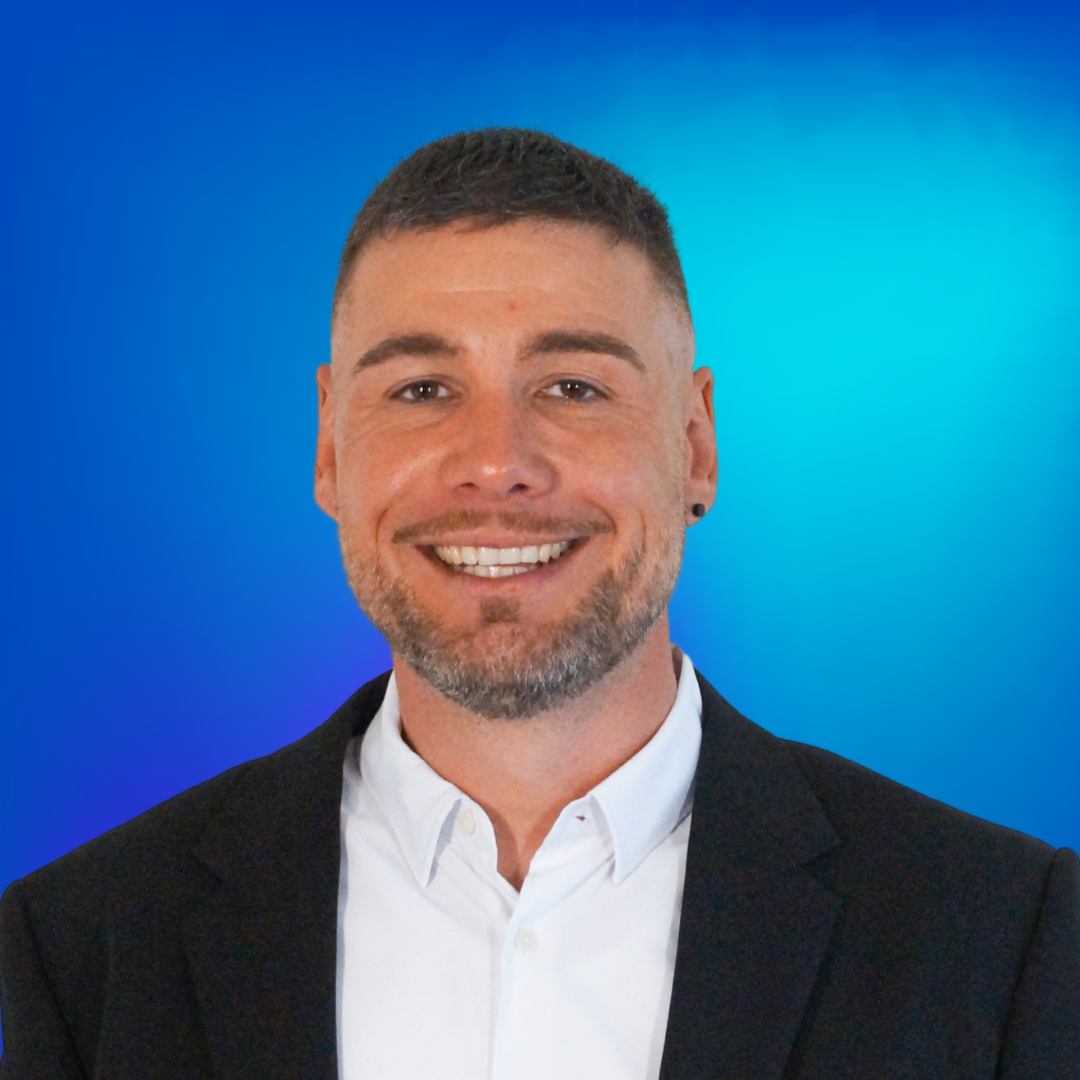 Porträt eines lächelnden Mannes mit Bart und kurzen dunklen Haaren, in weißem Hemd und schwarzem Sakko vor blauem Hintergrund.