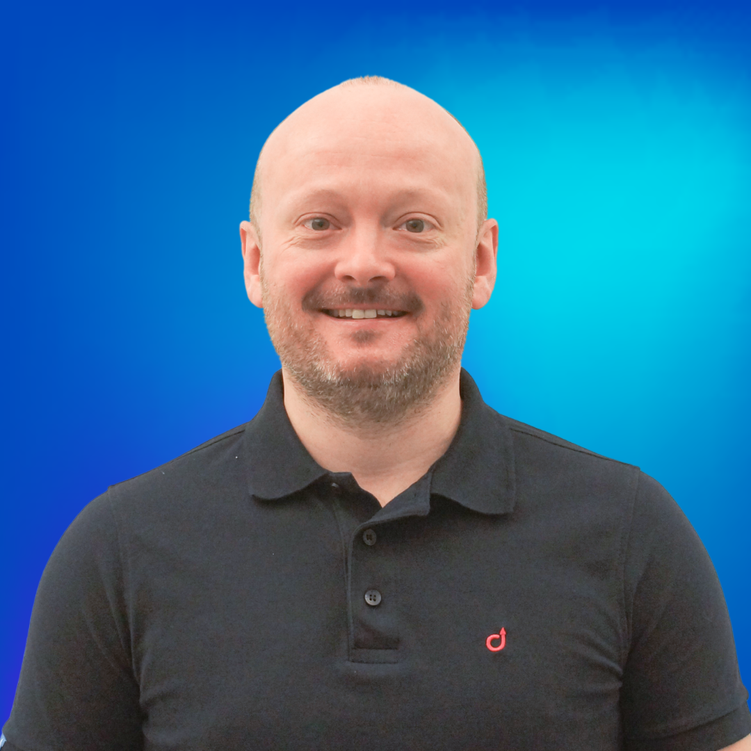 Ein bärtiger Mann lächelt in die Kamera, trägt ein dunkles Poloshirt vor blauem Hintergrund.