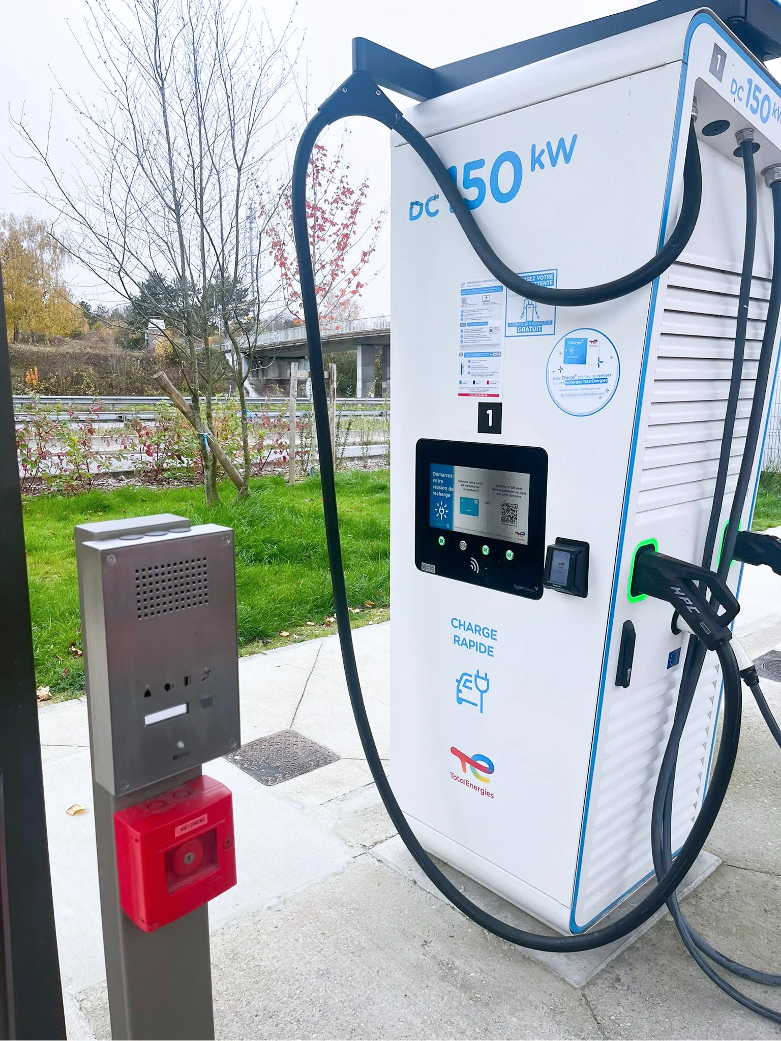 Eine TotalEnergies DC 150 kW Ladestation für Elektrofahrzeuge mit einer Sprechstelle und Notruf-Knopf für Sicherheit.