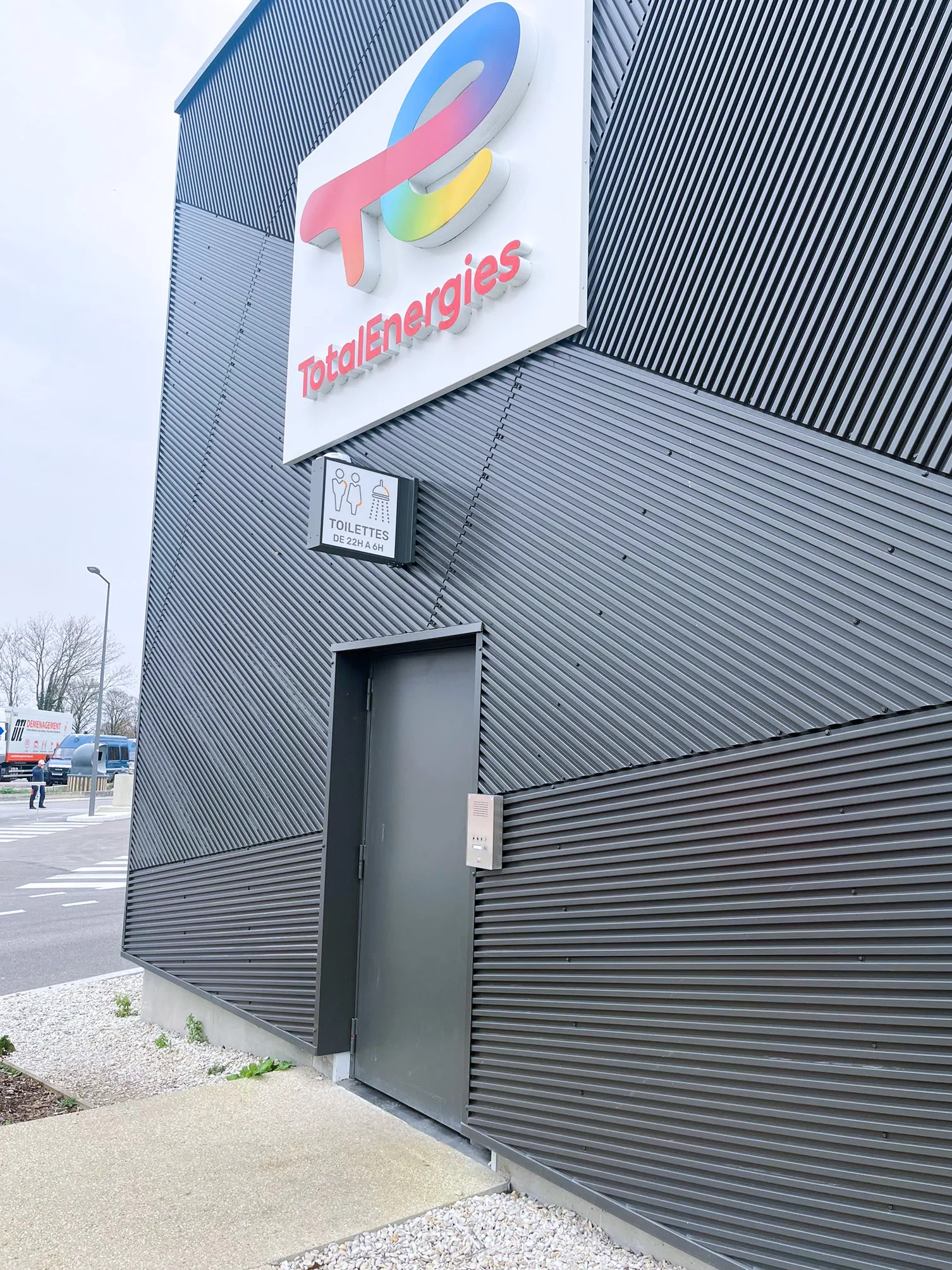 Eine moderne Wellblechfassade eines TotalEnergies Gebäudes. Eine Sprechanlage ist neben der Tür zu WC und Duschen angebracht.