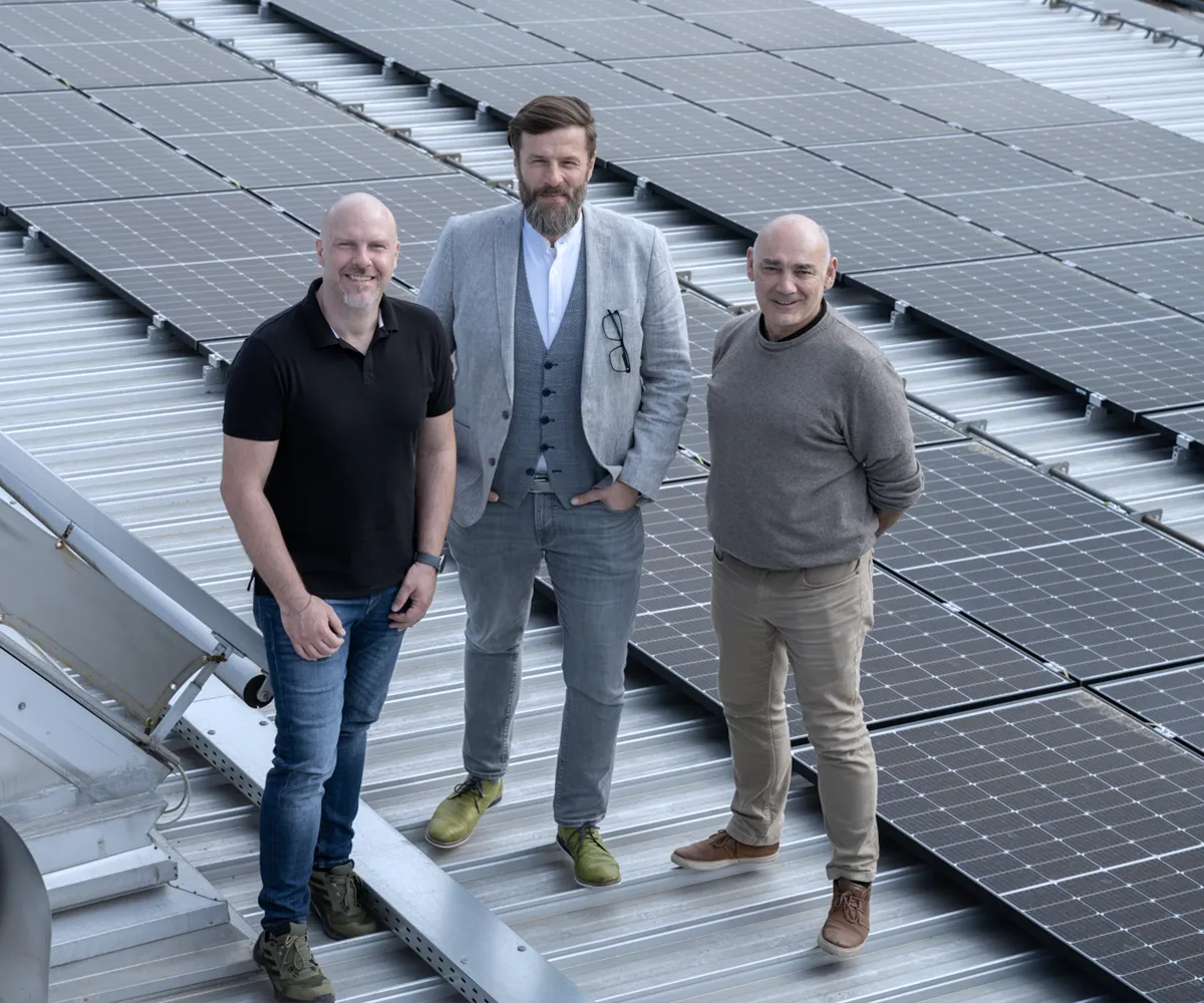 Geschäftspartner auf Solardach Drei Männer, einer davon mit Bart, stehen lächelnd auf einem sonnigen Dach mit vielen Solarmodulen.