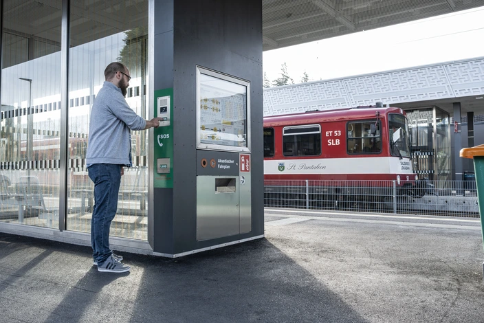 Mann an Sprechstelle im Bahnhof Mann an einem Bahnsteig drückt einen SOS-Knopf an einer Sprechanlage neben einem akustischen Fahrplan.