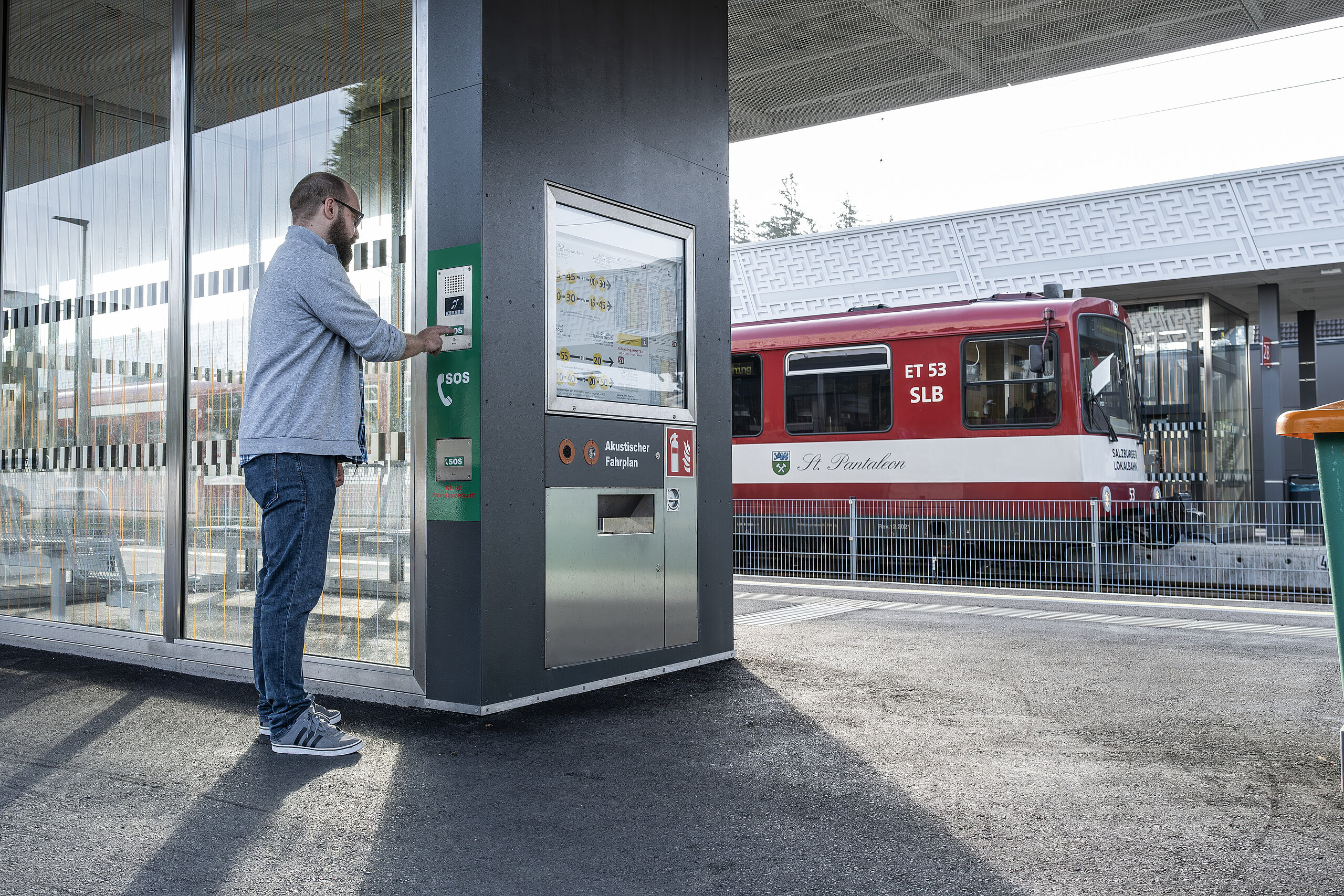 Mann an einem Bahnsteig drückt einen SOS-Knopf an einer Sprechanlage neben einem akustischen Fahrplan.