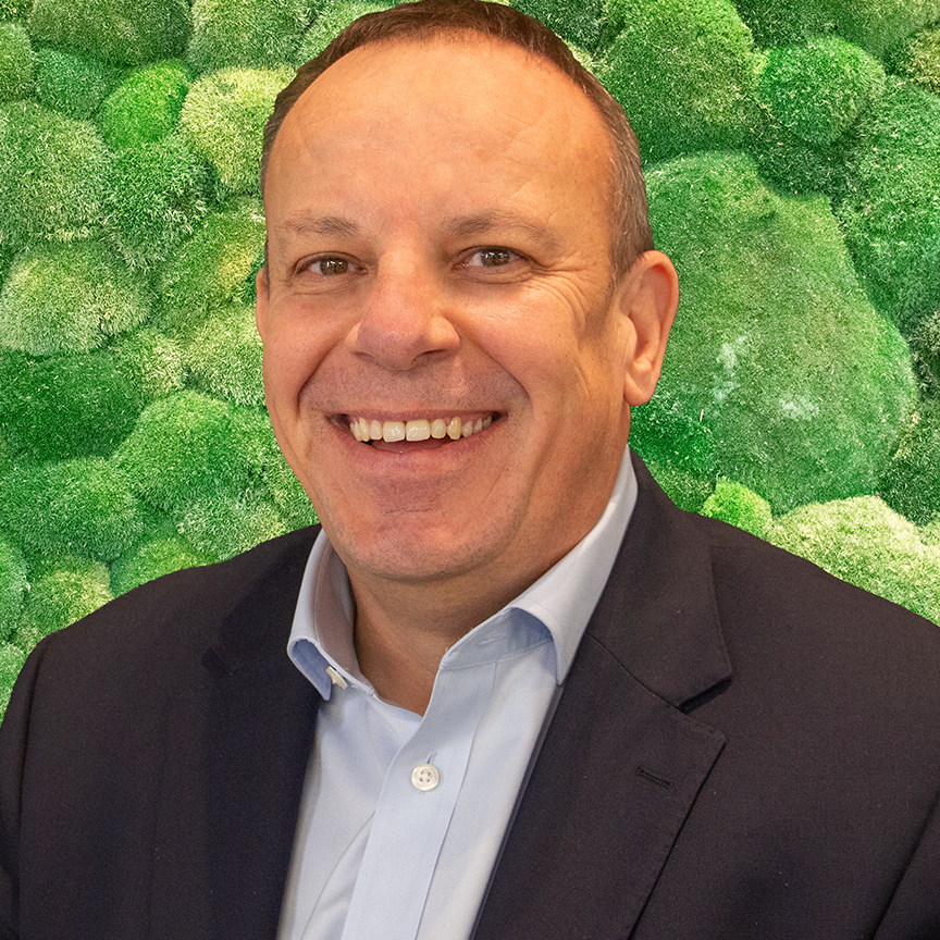 Smiling man in dark suit and light blue shirt against a green moss wall background. Professional headshot.