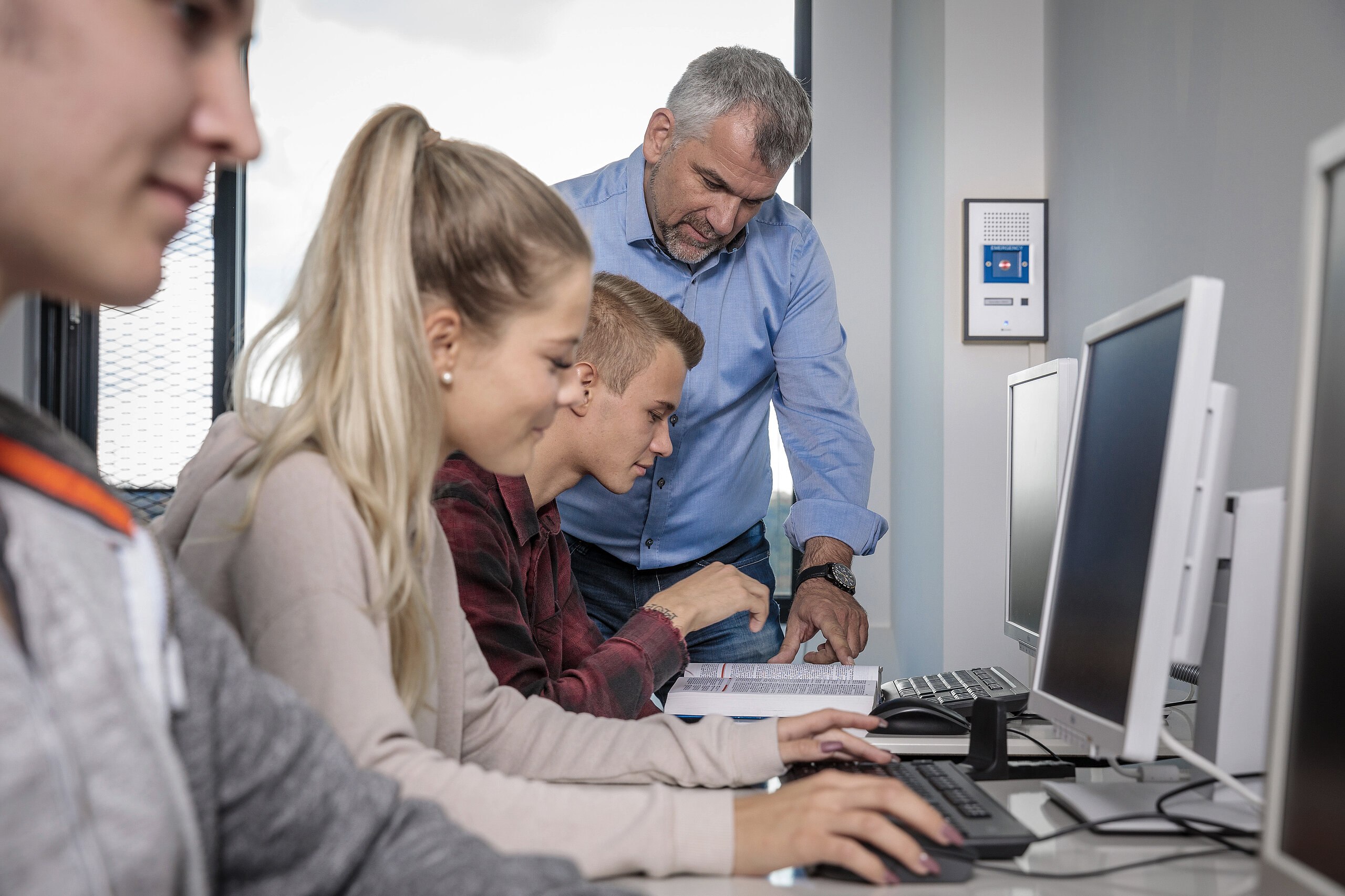 Lehrer hilft Schülern im Computerraum; Sprechanlage an der Wand.