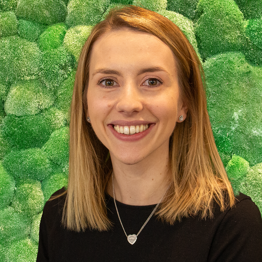 Headshot of a smiling young blonde woman in a black top against a vibrant green textured moss wall.