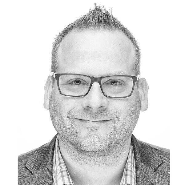 Headshot of a smiling man with glasses, spiky hair, stubble, plaid shirt, and blazer.