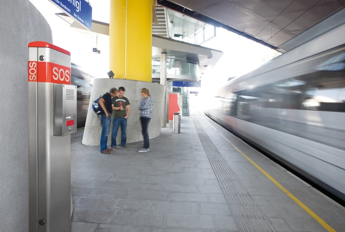 Notruf-Sprechanlage am Bahnsteig Silber-rote SOS-Sprechanlage auf einem Bahnsteig, mit unscharfem, vorbeifahrendem Zug und Menschen.