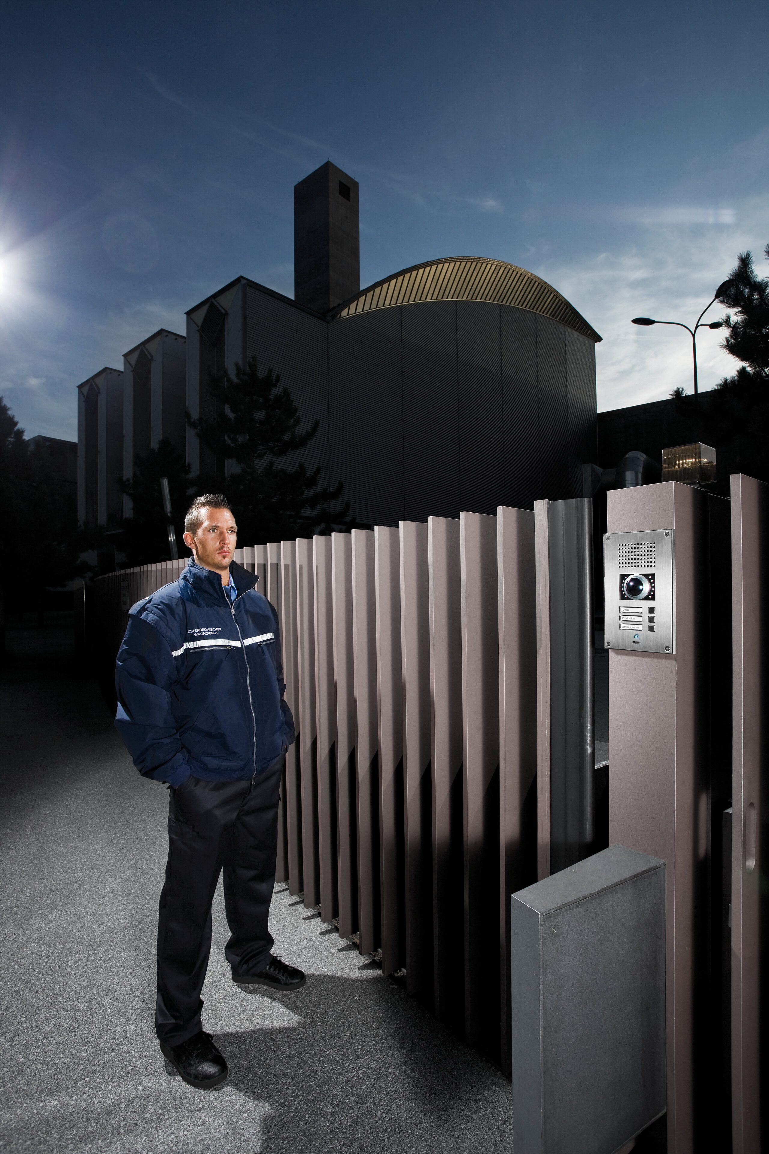 Uniformed security officer beside a modern gate with an intercom at a corporate building entrance.