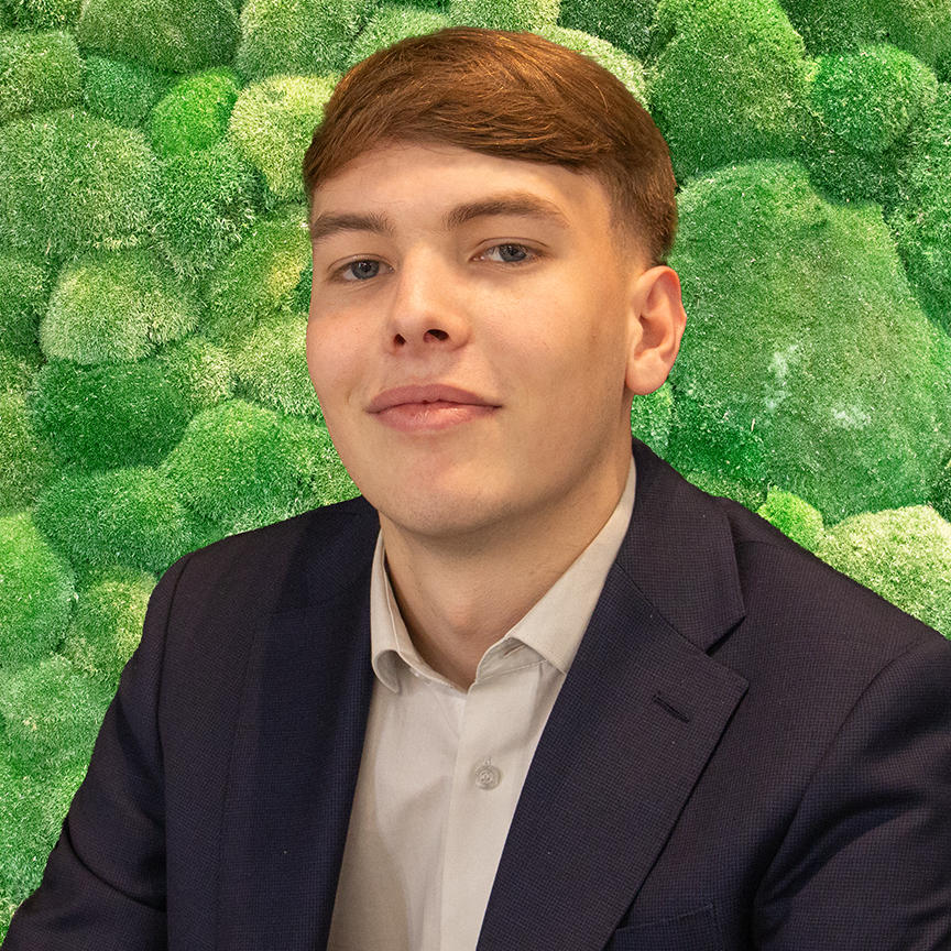 A young man in a blue suit smiles in front of a green moss wall.