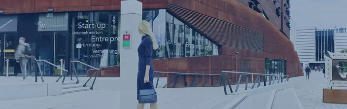Sprechanlage am modernen Campus Frau bei einer Sprechstelle vor einem rostfarbenen Universitätsgebäude.