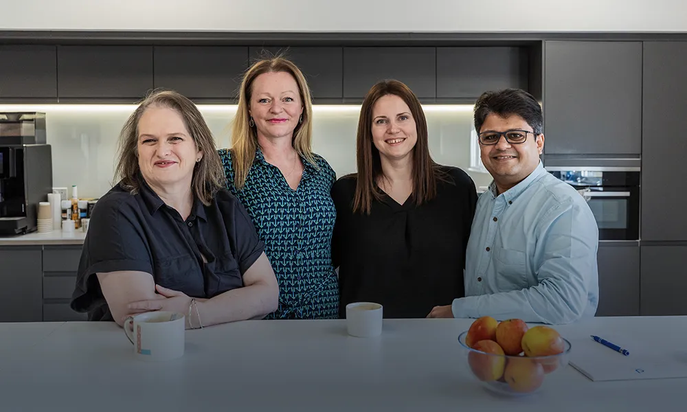 Four smiling colleagues in an office kitchen, standing behind a counter with mugs and apples.