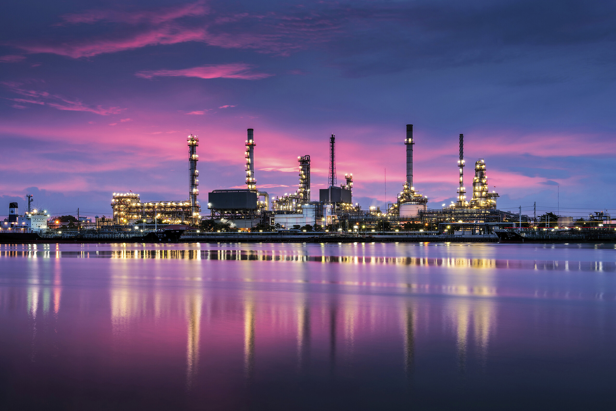 Industrial oil refinery glowing at night, reflected in water under a pink and purple sky.