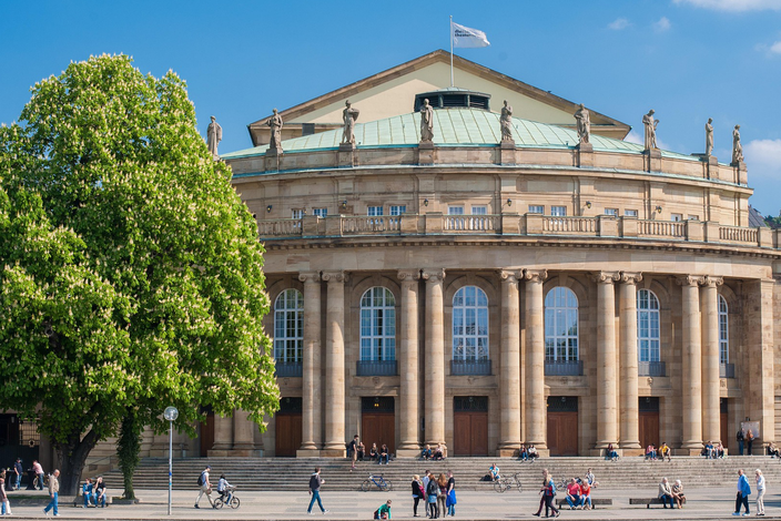 Staatstheater Stuttgart im Frühling Klassisches Theatergebäude mit Säulen, Statuen, blühendem Baum und Menschen an einem Sonnentag.