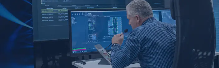 Operating a Communication System Man with microphone at control desk, viewing security system and floor plans on multiple monitors.