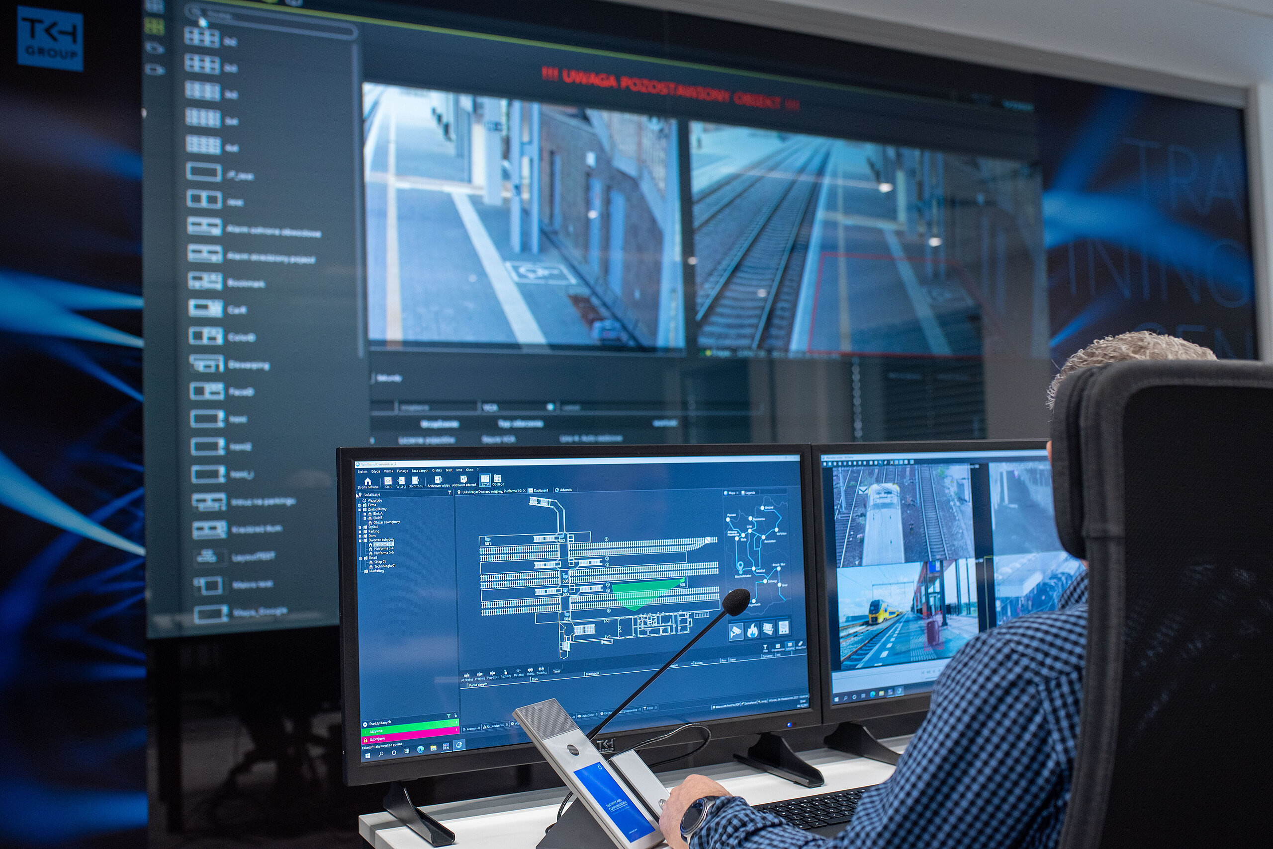 An operator in a control room views screens showing railway tracks, security camera feeds, and a communication system.