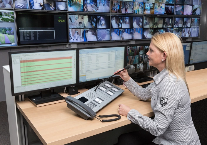 Operator using an intercom console with a gooseneck microphone in a security control room with many surveillance screens.