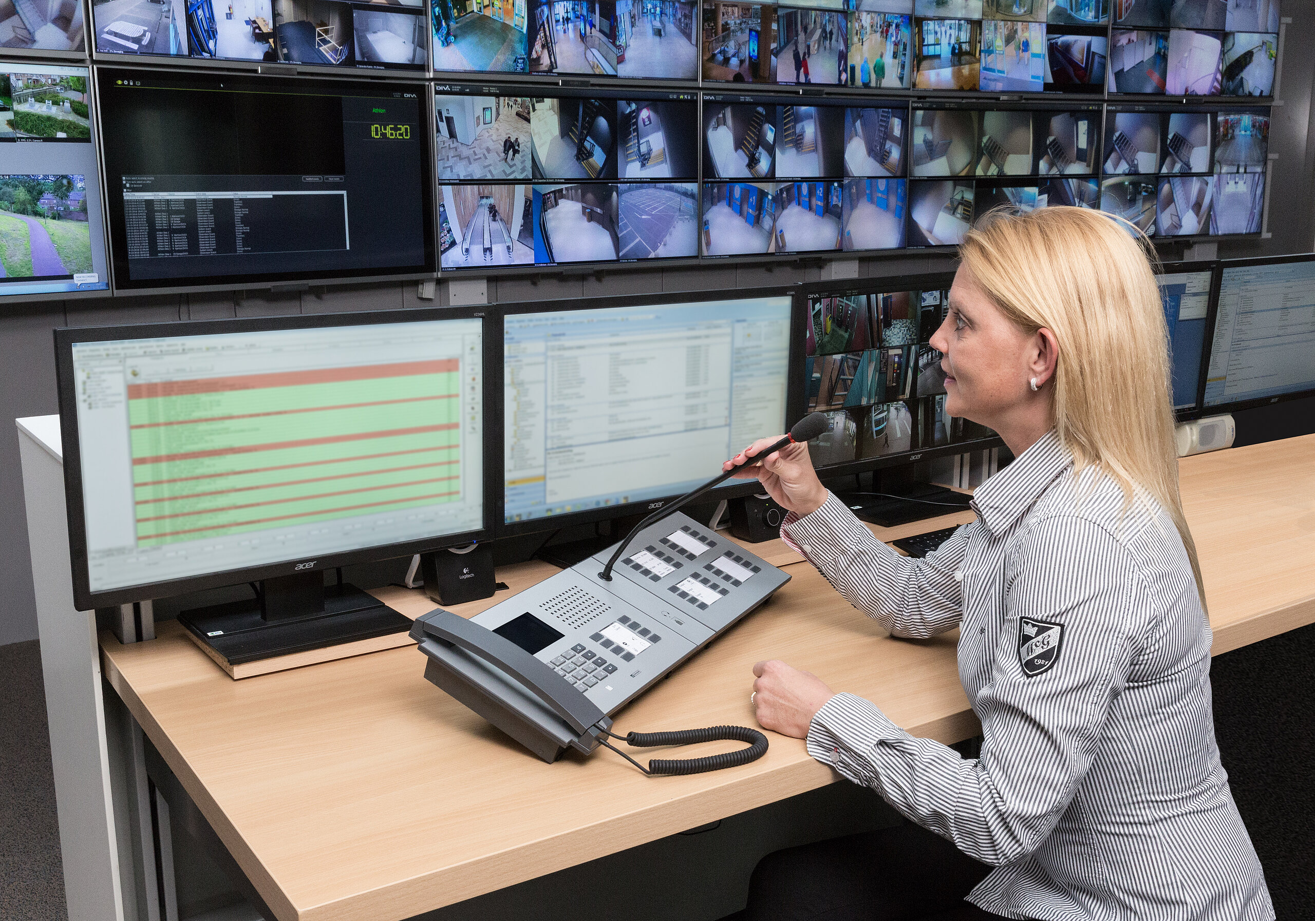 Bedienerin an einer Sprechanlage mit Schwanenhalsmikrofon in einem Überwachungsraum mit vielen Bildschirmen.